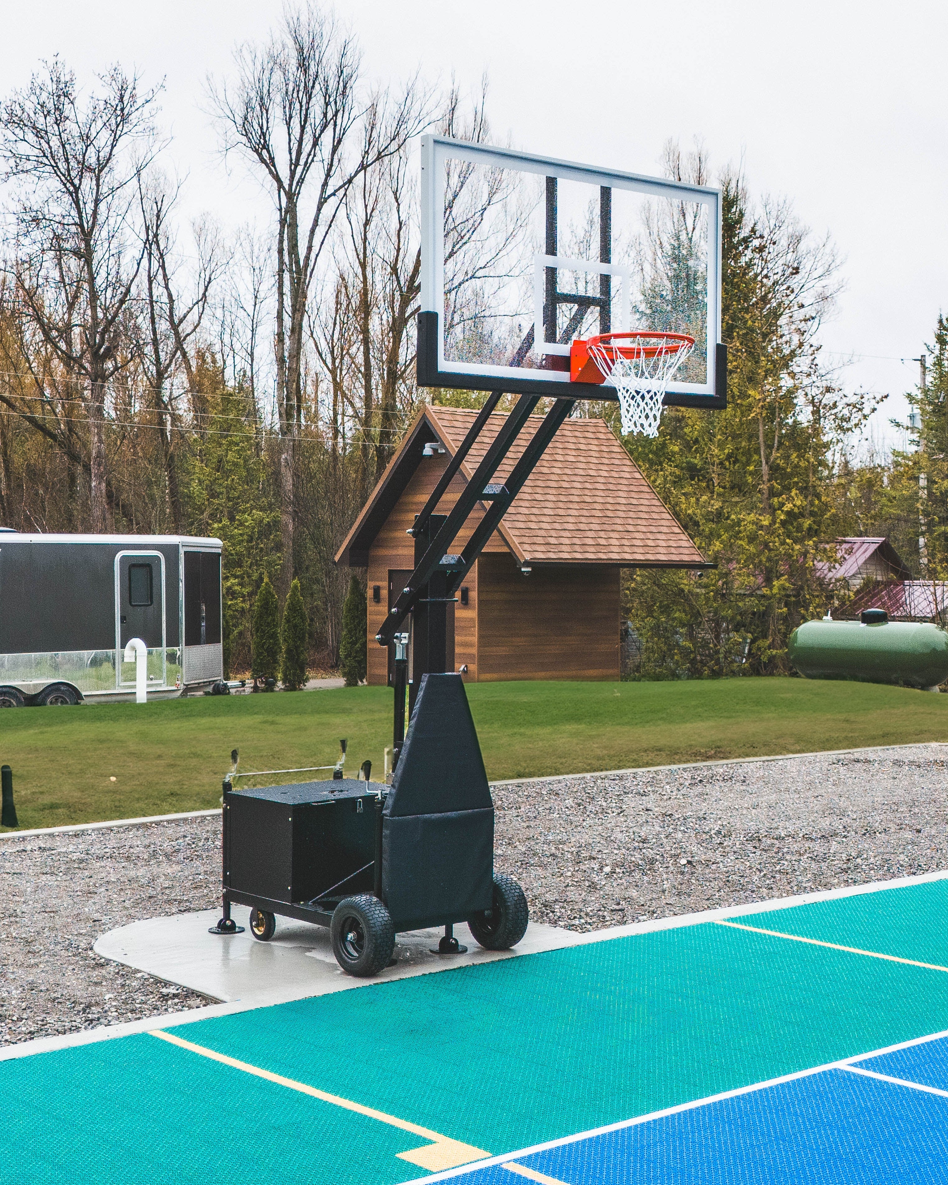 Outdoor basketball court with city hoops portable hoop system, trailer, and cabin in the background.