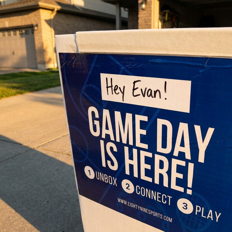 A white cardboard shipping box outdoors on a driveway, featuring a blue and white printed label that reads 'GAME DAY IS HERE!' with steps to unbox, connect, and play, for an eightynine sports hexpro sport tile court kit